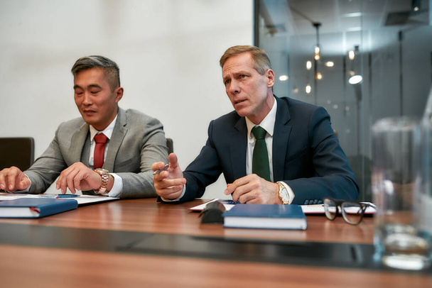 Business partners. Portrait of caucasian and asian business people in formal wear sitting at office desk. Multicultural team discussing project details at meeting - Foto, immagini