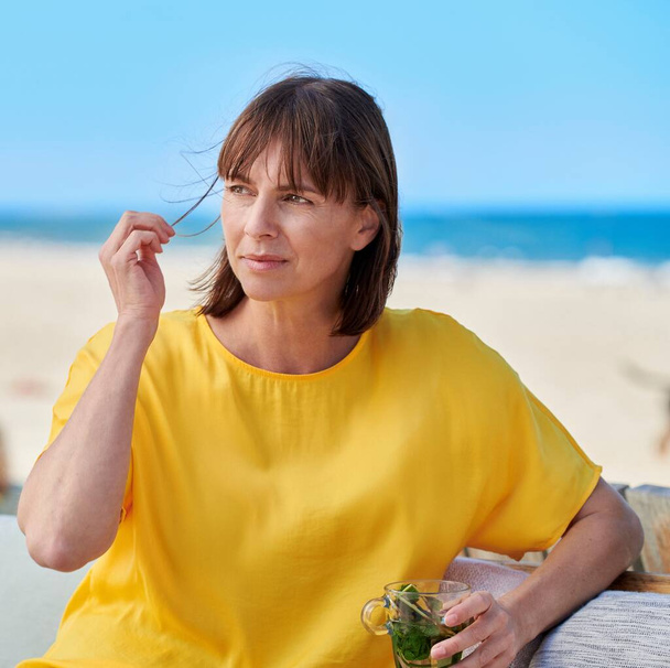 Mujer en la playa mirando preocupado infeliz amarillo blusa azul fondo viento jugando a través de su pelo soleado nublado tiempo
 - Foto, imagen