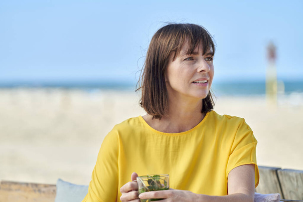 Kobieta na plaży patrząc zmartwiony nieszczęśliwy żółty bluzka niebieski tło wiatr grając koryta jej włosy słoneczne zachmurzenie pogoda - Zdjęcie, obraz