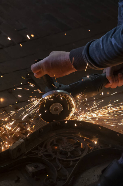Vue latérale des mains d'un homme travaillant sur une partie métallique d'un banc de jardin, à l'aide d'une meuleuse électrique pendant que des étincelles volent dans l'atelier industriel
. - Photo, image