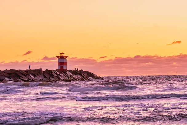 Красно-белый полосатый маяк во время яркого заката. Вода бьет по черным валунам волнами. Драматическое небо в золотом цвете. Шевенхель, Нидерланды
. - Фото, изображение
