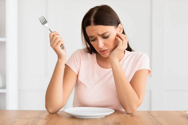 Голодная женщина смотрит на пустую тарелку, похудевшую дома
 - Фото, изображение