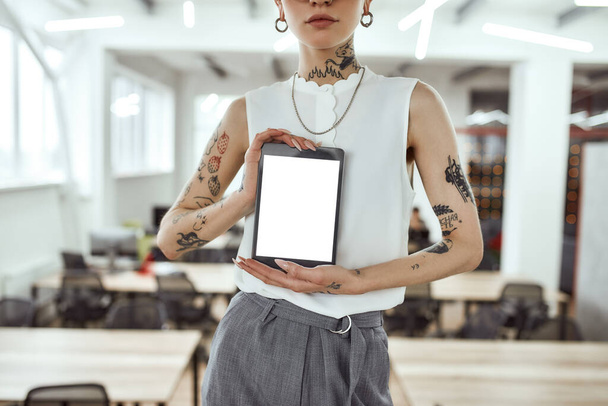 Showing new gadget. Cropped photo of tattooed business woman holding digital tablet while standing in modern coworking space - Foto, afbeelding