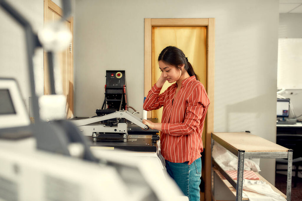 Quality printing. Young cheerful female worker using heat press transfer machine for printing advertisement sticker with text on t-shirt at workplace - Valokuva, kuva
