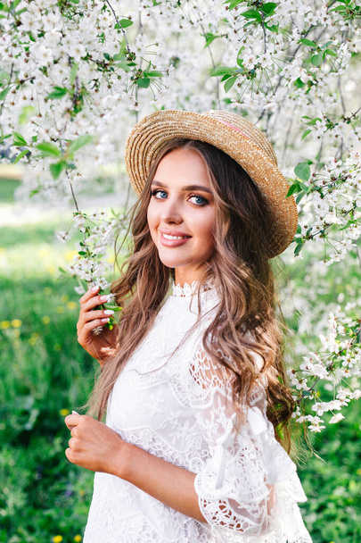 young attractive woman with curly long hair in straw hat enjoying blooming garden in sunny spring day. - Фото, изображение