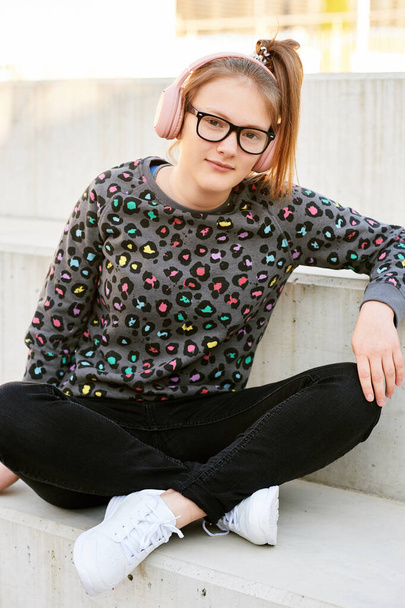 Ritratto all'aperto di una giovane adolescente che si riposa sui gradini accanto alla scuola, indossando felpa con stampa animalier, jeans neri e sneakers bianche
 - Foto, immagini
