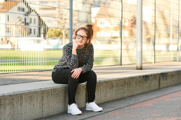 Retrato al aire libre de una joven adolescente que descansa en los escalones junto a la escuela, con sudadera con estampado de animales, jeans negros y zapatillas blancas.
 - Foto, Imagen