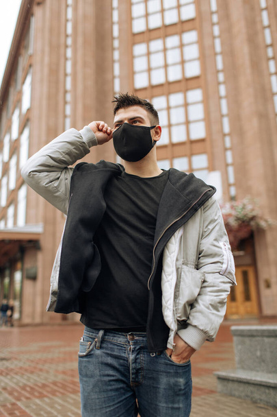 Brunette guy in a black tracksuit puts on a face mask. Black stylish face mask during a pandemic virus crown. Handmade cotton face mask for protection against viruses and bacteria. - Valokuva, kuva
