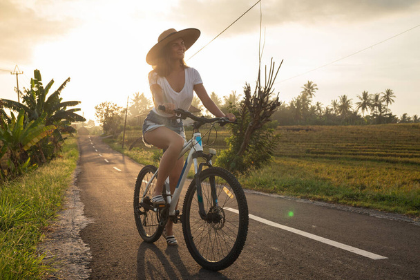 Young happy woman is riding bicycle by narrow country road - Valokuva, kuva