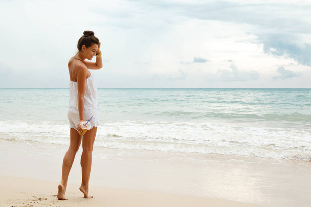 Красивая женщина в белом комбинезоне гуляет по пляжу с бокалом ледяного кофе
  - Фото, изображение