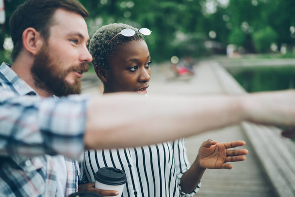 Bearded guy pointing away and explaining route to African American lady with drink to go on blurred background of park - 写真・画像