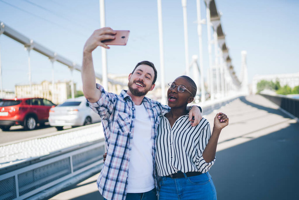 Smiling excited bearded guy calling friend on video chat hugging African American content young woman in glasses with short hair on bridge - Foto, immagini
