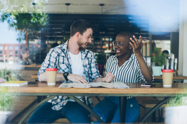 Cheerful casual multiracial couple talk, discuss plans for summer trip, preparing for vacation, smiling while sitting at table at cafe - Foto, afbeelding