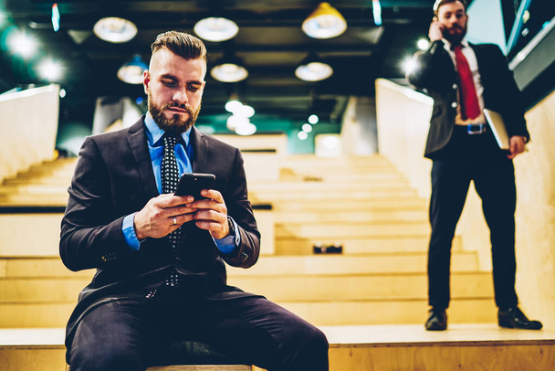 Elegant serious man in formal suit relaxing from work using mobile chatting with friend sitting on stairs of modern building - Photo, image