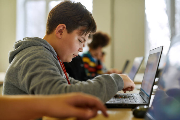 Learners Today, Leaders Tomorrow. Close up of caucasian schoolboy looking at the screen of the laptop together with other pupils during a lesson in modern smart school - Fotografie, Obrázek