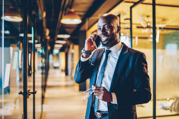 Happy confident African American male manager in suit looking away and talking on cellphone while holding glasses and standing in modern glass office corridor - Photo, image
