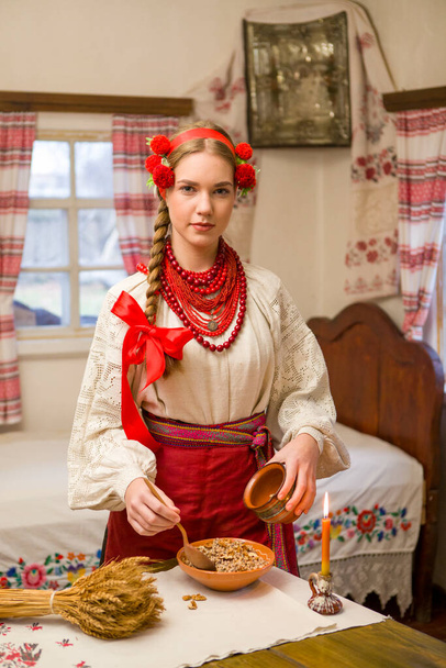 Ulusal elbiseli güzel bir kız şenlikli bir akşam yemeği hazırlıyor. Güzel bir çelenk ve kırmızı işlemeli bir elbise içinde. Aile kutlaması ve ulusal geleneğin kutlanması. Kutia ile kase - geleneksel - Fotoğraf, Görsel