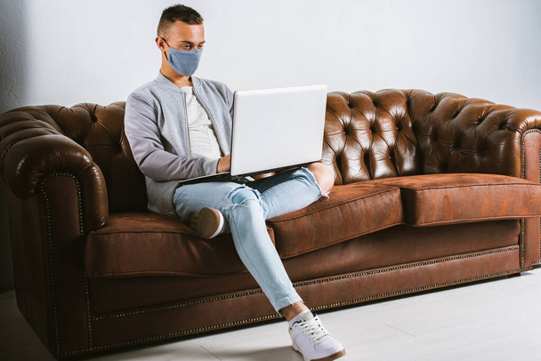 Gri maskeli genç bir adam koronavirüsün yayılmasını önlemek için karantina sırasında dizüstü bilgisayarıyla uzaktan çalışıyor. Adam COVID-19 salgını sırasında evden çalışıyormuş. - Fotoğraf, Görsel