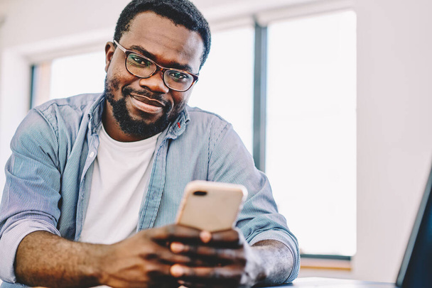 Afroamericano sonriente barbudo optimista hombre en gafas de texto en el teléfono inteligente con placer durante el descanso de trabajo en interiores mirando a la cámara - Foto, imagen