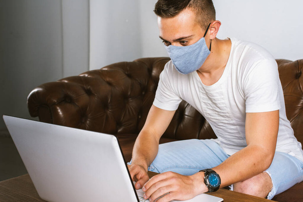 Um jovem com uma máscara facial cinza trabalha remotamente em seu laptop durante a quarentena para evitar a propagação do coronavírus. O cara trabalha em casa durante a pandemia do COVID-19
.  - Foto, Imagem