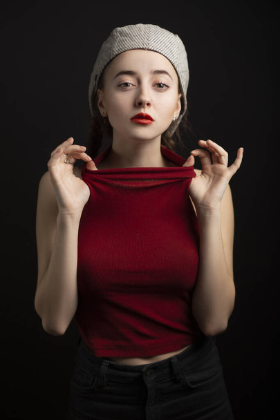 Cunning look of a girl with red lips on a chair on a black background in a cap - Photo, Image