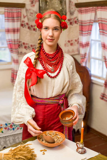 Kaunis tyttö kansallispuvussa valmistelee juhlapäivällistä. Kaunis seppele ja punainen kirjailtu mekko. Perhejuhla ja kansallisen tavan juhliminen. Bowl kanssa Kutia - perinteinen - Valokuva, kuva
