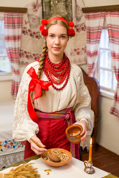 Kaunis tyttö kansallispuvussa valmistelee juhlapäivällistä. Kaunis seppele ja punainen kirjailtu mekko. Perhejuhla ja kansallisen tavan juhliminen. Bowl kanssa Kutia - perinteinen - Valokuva, kuva