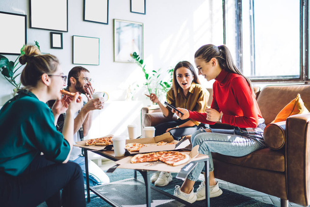 Cheerful woman in casual outfit browsing smartphone while smiling people eating delivered pizza and sitting on couch by table in stylish apartment - Foto, Bild