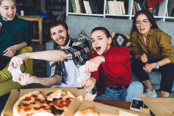 Focused screaming friends in comfortable casual wear enjoying video game while chilling near sofa and eating pizza in trendy domicile - Фото, изображение