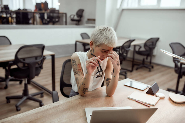 Feeling frustrated. Tired young tattooed business woman keeping eyes closed, holding her glasses in hand and massaging nose while sitting at her working place in the modern office - Zdjęcie, obraz
