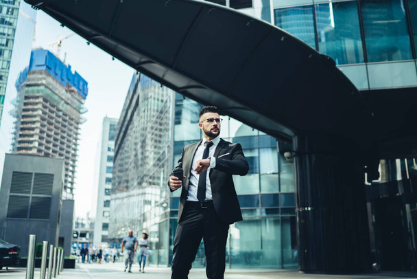 Serious hurrying stylish male employee in corporate suit with coffee to go thinking about time on wrist watch in city center looking away - Foto, Imagen
