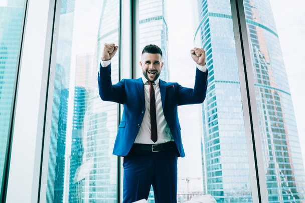 Happy smiling male with arms up in formal suit with red tie amazed by success of startup looking at camera at workplace - Foto, Imagem