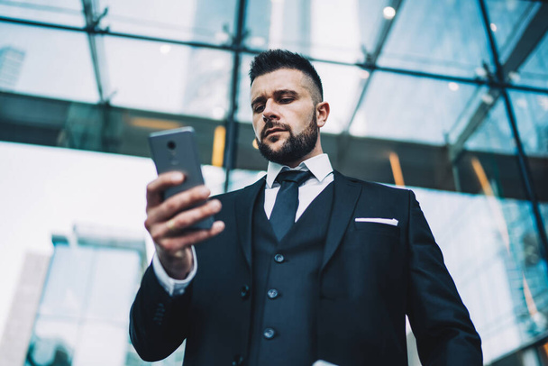 From below focused businessman in suit frowning and browsing smartphone while standing outside contemporary building in downtown of modern city - Foto, immagini