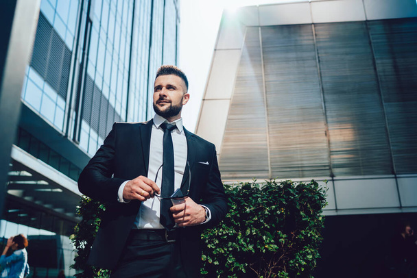 Low angle of confident boss in dark suit carrying sunglasses and smartphone and looking away while standing near shrub outside modern buildings on city street - Fotografie, Obrázek