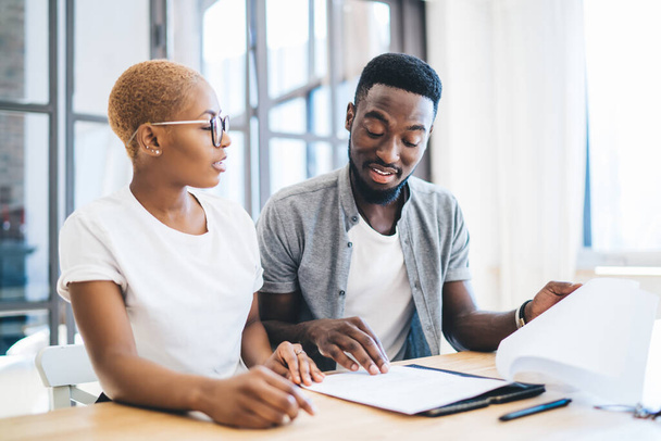 Focused black couple sitting at table in apartment in front of realtor reading estate contract with attention and talking with concentration - Foto, Bild