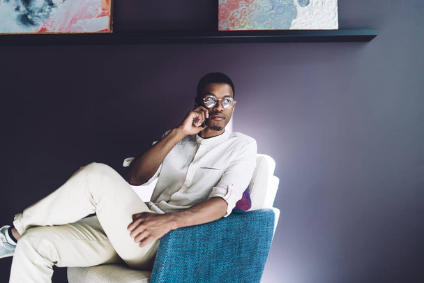 African American stylish man in trendy clothes speaking on smartphone thoughtfully while sitting and lounging on chair on purple wall background in cafe - Foto, Imagem
