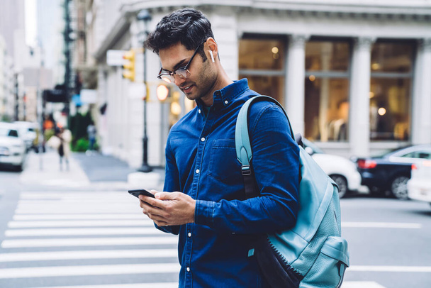 Focused handsome ethnic adult man in casual shirt and glasses browsing on smartphone while standing on busy city street in daylight - Foto, imagen