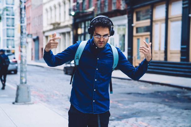 Energetic modern ethnic adult man in earphones and eyeglasses with backpack moving hands while dancing on New York city street  - Фото, изображение