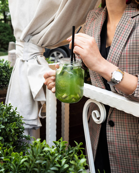 Cold kiwi lemonade and lime. Mug in the form of a jar. Drink with ice, green color. Against a background of flowers and greenery. The girl is holding a cocktail in her hand. Menu for bars, cafes and restaurants. Vegetarian. - Photo, image