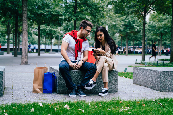Sweet couple chilling on stone bench near paper bags after shopping and using smartphone outside in park on blurred background - Fotografie, Obrázek
