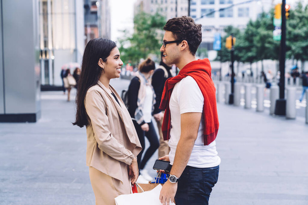 Side view of loving diverse smiling couple in casual outfit speaking on street with shopping bags while using smartphone and looking at each other on blurred background - Foto, Imagem