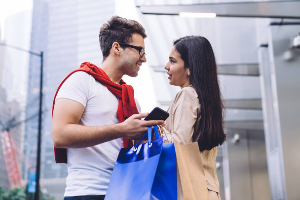 Low angle side view of loving multiracial couple in casual wear while using smartphone and resting near urban building with shopping bags and looking at each other on blurred background - Foto, Bild