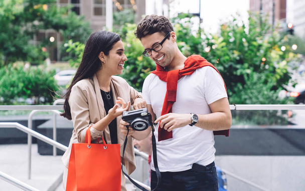 Smiling content multiethnic couple with shopping bag resting on megapolis street while viewing photo camera in daylight on urban blurred background - Foto, afbeelding