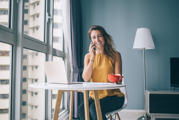 Portrait of happy female holding cup with coffee communicating with friend via cellphone app and smiling at camera, positive cheerful woman using home connection for making international call - 写真・画像
