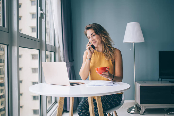 Satisfied young woman in yellow sleeveless blouse sitting at white round table with laptop and red mug communicating on smartphone at home - 写真・画像