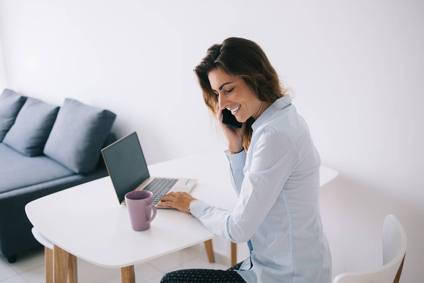 Side view of young smiling brunette sitting near cup and laptop with blank space on white table while talking on phone - Φωτογραφία, εικόνα