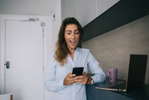 Beautiful brunette woman with open mouth excited and shocked while reading unexpected great news on smartphone and standing at home near laptop and cup - Foto, immagini