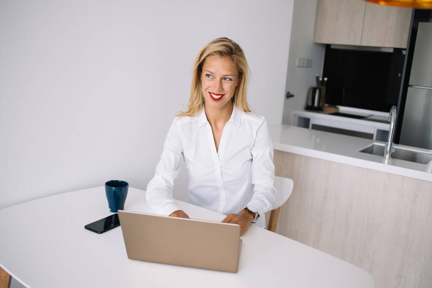 Young blonde in white shirt sitting at desk and smiling while working at laptop and looking away in office against white wall  - Foto, Imagem