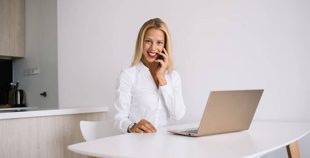 Delighted young blonde woman sitting at white table in blouse speaking on smartphone using laptop at home and looking at camera - Foto, immagini