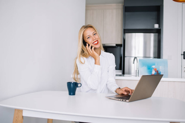 Positive young blonde woman sitting at white table with blue cup in blouse speaking on smartphone using laptop at home and looking at camera - Fotó, kép
